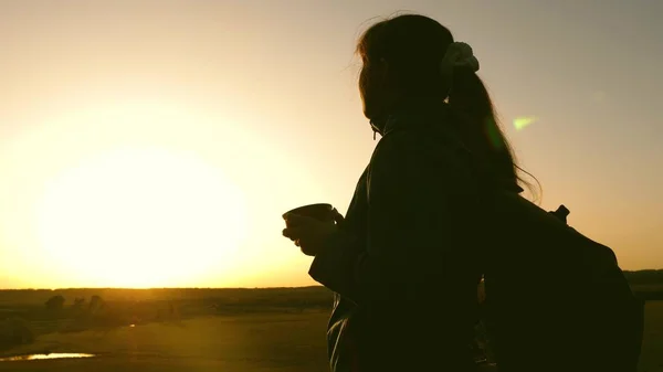 Viaggiatore donna silhouette, si erge sulla cima di una collina bevendo caffè in vetro da thermos. ragazza turistica beve tè caldo e guarda il sole. Avventuriero riposo dopo aver raggiunto l'obiettivo. concetto di libertà . — Foto Stock