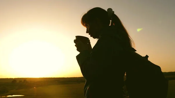 Силуэт женщина путешественник, стоит на вершине холма пить кофе в стекле из термоса. Туристка пьет горячий чай и смотрит на солнце. Авантюрист отдыхает после достижения цели. свобода концепция . — стоковое фото