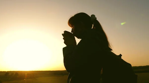 Silhouette Frau Reisende, steht auf einem Hügel und trinkt Kaffee in Glas aus Thermoskannen. Touristenmädchen trinkt heißen Tee und blickt in die Sonne. Abenteurer ruhen nach dem Erreichen des Ziels. Freiheitskonzept. — Stockfoto