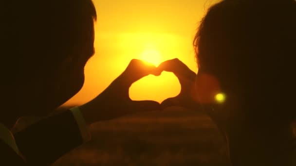 Een man en een vrouw tonen een hart met hun handen bij de zonsondergang van de gouden zon. Liefhebbers van een romantische reis. Teamwork van een liefdevolle paar. Het concept van een gelukkige familie. Close-up. — Stockvideo