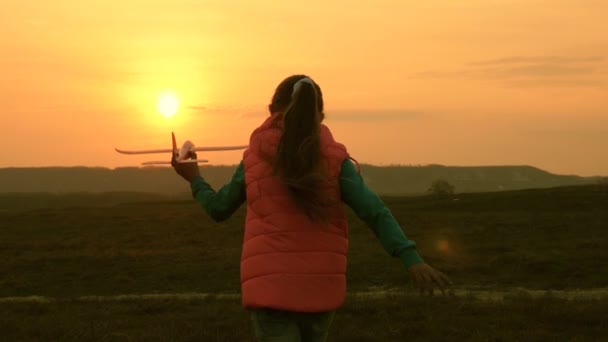 Uma criança joga um avião de brinquedo. menina sonha em voar e se tornar um piloto. Movimento lento. Menina feliz correndo com um avião de brinquedo no fundo de um pôr do sol sobre o campo. conceito de uma família feliz . — Vídeo de Stock