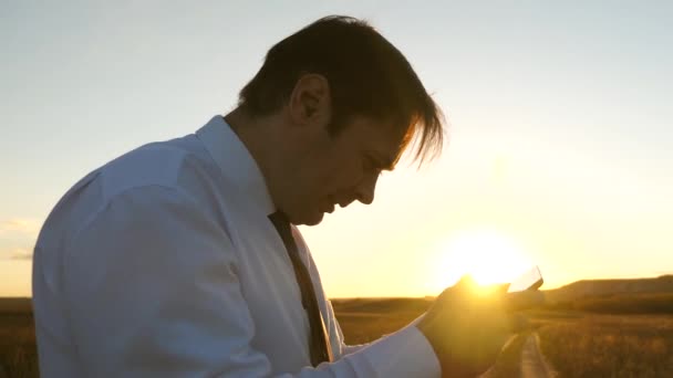 Geschäftsmann, der im Park in den warmen Strahlen des Sonnenuntergangs Tablet-Spiele spielt. Mann spielt Spiele auf dem Smartphone. glücklicher Mann in weißem Hemd und Krawatte spielt online auf Tablet. — Stockvideo