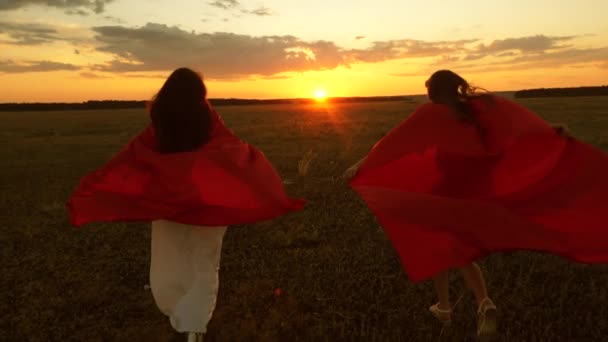 赤いマントの女の子は、日没時にスーパーヒーローを再生します。チームワーク。ガールフレンドは夕日の夕方のフィールドを横切って実行します. — ストック動画