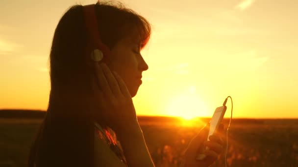 아름 다운 석양의 광선에 음악과 춤을 듣고 행복 한 소녀. 헤드폰의 어린 소녀와 스마트 폰이 태블릿의 센서에 손가락을 터치 하 여 온라인으로 노래를 선택 합니다. 슬로우 모션. — 비디오