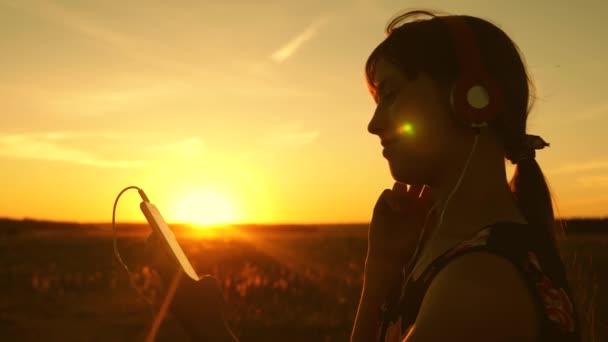 Fille heureuse écoutant de la musique sur smartphone dans les rayons d'un beau coucher de soleil de printemps. Au ralenti. jeune fille avec écouteurs et avec tablette sélectionne des chansons en ligne, dans le parc en été dans les rayons de soleil . — Video