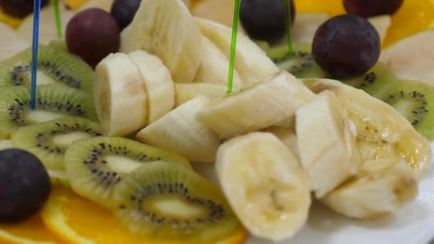 BanaJag, apelsiner, vindruvor, kiwi skivad, närbild. Färsk frukt maträtt på en festlig matbord. Diverse skivad frukt spett på en tallrik. — Stockvideo