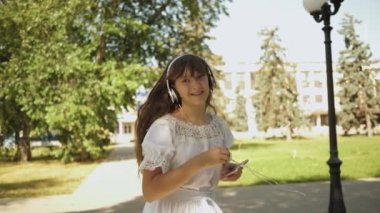 kız kulaklıkla telefonla seyahat eder, müzik dinler, gülümser. neşeli kız dansları ilkbaharda yaz aylarında parka gider. güzel kız dans, elini sallayarak ve seyahat için sizinle gitmek için arıyor.