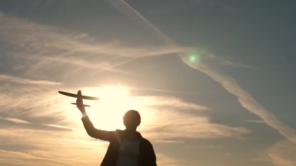 Девушка играет с игрушечным самолетом на закате. Мечтает летать. Счастливое детство. Дети на фоне солнца с самолетом в руке. Силуэт детей, играющих в самолете — стоковое видео
