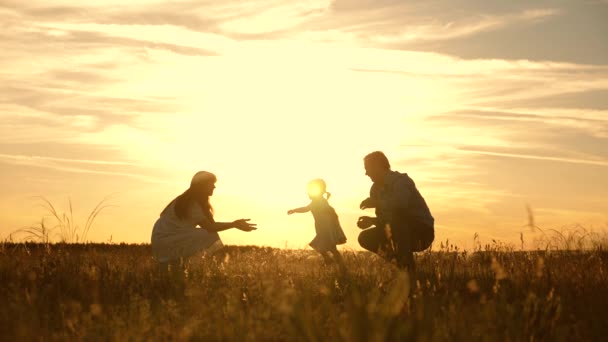 Família feliz brincando com sua filha nos raios do sol. Mãe e pai brincando com a criança no parque ao pôr do sol, criança dá os primeiros passos. criança vai de pai para mãe e ri. Movimento lento . — Vídeo de Stock