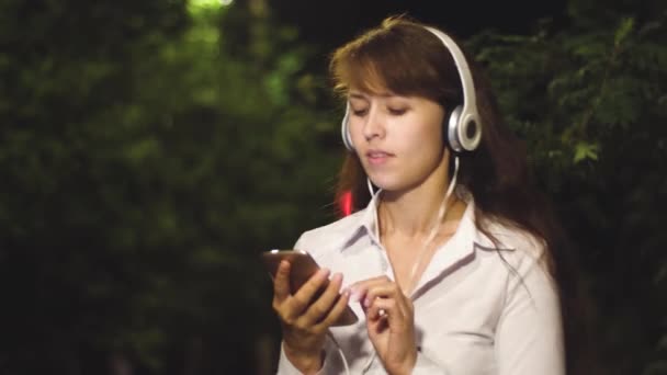 Веселая девушка путешествует по ночному городу. Молодая девушка в наушниках и смартфонах слушает музыку, танцует и улыбается ночью в городском парке. макро — стоковое видео