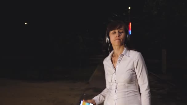 Niña en los auriculares y el teléfono inteligente está escuchando música, bailando y sonriendo por la noche en un parque de la ciudad. alegre chica viaja en la noche de la ciudad . — Vídeo de stock