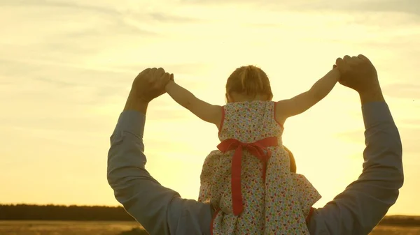 Отец ходит с дочерью на плечах в лучах заката. Папа носит на плечах своего любимого ребенка, переходя поле. Ребенок с родителями гуляет вечером на закате. Медленное движение . — стоковое фото