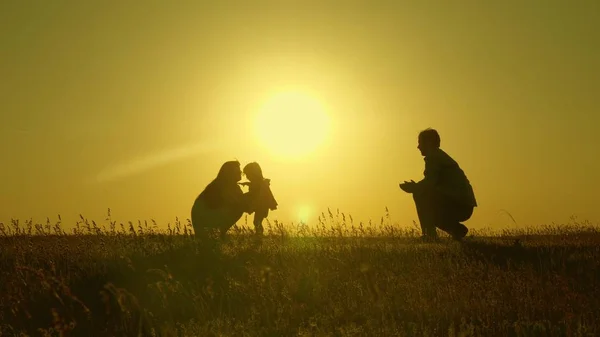 Mamma och pappa leker med sin dotter i solen. Happy baby går från pappa till mamma. ung familj på fältet med ett barn 1 år. familj Happiness Concept. vackert solsken, solnedgång. — Stockfoto
