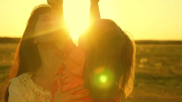Madre felice cammina con la figlia tra le braccia nei raggi del tramonto. La mamma sta parlando con il bambino felice, il bambino è seduto sulle braccia delle madri al tramonto del sole dorato. Rallentatore . — Foto Stock