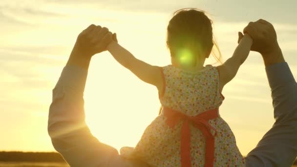 お父さんは、太陽の下で娘と肩の上で踊っています。父は日没の光の中で赤ん坊を肩に乗せ、旅をする。両親を持つ子供は日没時に歩きます。公園で休む幸せな家族。家族の概念 — ストック動画