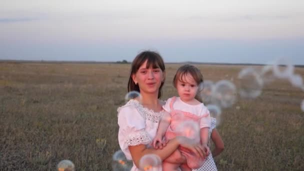 Mère heureuse jouant avec des enfants soufflant des bulles de savon. les enfants attrapent des bulles et rient dans le parc en été. Au ralenti. Le concept d'une famille heureuse. enfant, soeurs et mère jouent ensemble . — Video