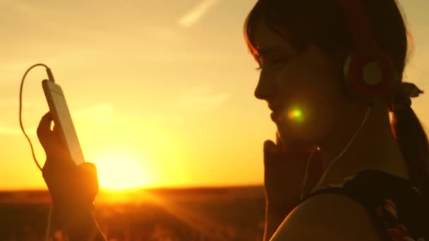 Menina viajante ouvindo música no smartphone em raios de um belo pôr do sol de primavera. jovem com fones de ouvido e com tablet seleciona músicas on-line, no parque no verão em raios de sol . — Vídeo de Stock