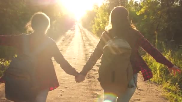 Hiker Girl. happy girls travelers with backpacks run along country road holding hands in rays of the bright sun. concept of sports tourism and travel. children travelers. teen girls travel and hold — Stock Video