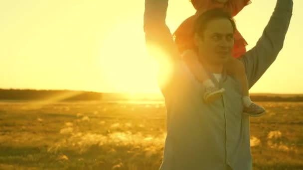 Father plays with his daughter on his shoulders in the rays of sunset. Dad carries on shoulders of his beloved child, walking across field. A baby with parents walks in evening at sunset. Slow motion. — Stock Video