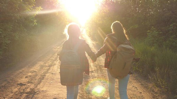 Hiker Girl. happy girls travelers with backpacks run along country road holding hands in rays of the bright sun. concept of sports tourism and travel. children travelers. teen girls travel and hold