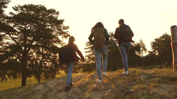 Três meninas viajam, caminham através de bosques para escalar colina se alegrar e levantar as mãos para o topo. Caminhante. meninas viajam com mochilas na estrada do campo. Família feliz em viagens de férias . — Fotografia de Stock
