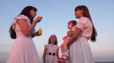 Kızları ve annesi gün batımında parkta baloncuklar üflüyor. Yavaş çekim. Mutlu anne sabun kabarcıkları üfleme çocuklarla oynarken. mutlu aile konsepti. bebek, kız kardeşi ve anne güneşte oynuyor.