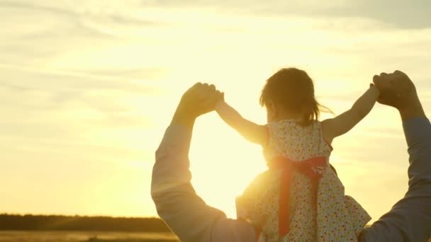 両親と一緒に赤ちゃんは夕暮れ時に夕方に歩きます。スローモーション。父は日没の日の光の中で娘と肩を並べて歩く。お父さんは、フィールドを横切って歩いて、彼の最愛の子供の肩を運びます. — ストック動画