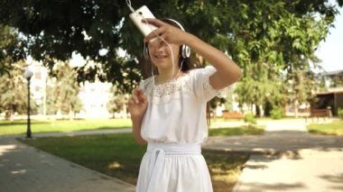 Peşimden gel, kız şehri dolaşıyor. Yavaş çekim. bir sırt çantası ile kız kulaklık park gider ve müzik ve gülümsüyor dinler, genç mutlu kamera lensi elini dalgalar.