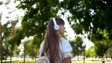bir sırt çantası ile kız kulaklıklar Park gider ve müzik dinler ve gülümsüyor, Genç mutlu kamera objektifi elini dalgaları. uzun saçlı beyaz bir elbise içinde kız şehir etrafında yolculuk. Yavaş hareket.