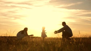 anne ve baba gün batımında parkta çocukla oynarken, çocuk ilk adımları atar. mutlu aile güneş ışınlarında kızları ile oynarken. çocuk babadan anneye geçer ve güler. Yavaş çekim.