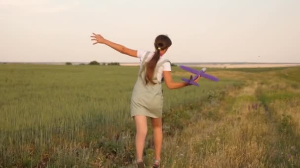 美しい女の子は小麦畑でおもちゃの飛行機で走ります。子供たちはおもちゃの飛行機をします。ティーンエイジャーは飛行し、パイロットになることを夢見ています。女の子はパイロットと宇宙飛行士になりたい. — ストック動画