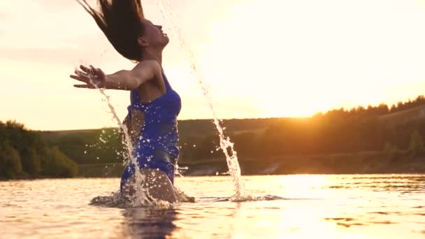 Игривая девушка брызгает своими длинными волосами в освежающий летний вечер возле тропического острова с освежающей речной водой. Девушка переворачивает волосы назад. красивый брызг воды в лучах заката. Всплеск воды — стоковое видео