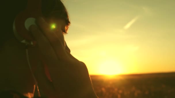 Молодая девушка мечтает под музыку на солнце. Девочка-подросток слушает музыку и смотрит на закат. Счастливая девушка танцует в наушниках в лучах прекрасного восхода солнца в парке . — стоковое видео