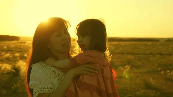 Šťastná matka chodí se svou dcerou do náručí v paprscích zapadajícího slunce. Máma mluví se šťastným dítětem, dítě sedí na matkách při západu slunce zlatého slunce. Zpomaleně. — Stock fotografie