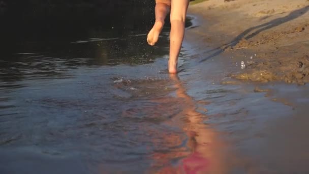 Dziewczyna biegnie boso wzdłuż brzegu i grając w wodzie na plaży o zachodzie słońca. rozprysków wody latać w różnych kierunkach. Teen Girl zabawy w lecie na plaży. Jasne słońce. Zwolnionym. — Wideo stockowe