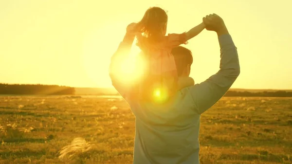 Papà danzava sulle spalle con sua figlia al sole. Papà viaggia con il bambino sulle spalle in raggi di tramonto. Un bambino con i genitori cammina al tramonto. famiglia felice che riposa nel parco. concetto di famiglia — Foto Stock