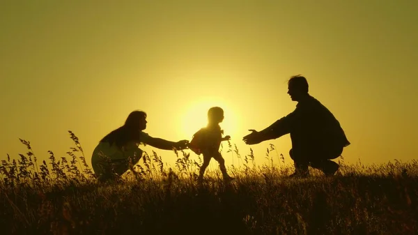 Hijita con padres saltando al atardecer. Siluetas de mamá papá y bebé en los rayos del amanecer. Concepto familiar. Caminar con un niño pequeño en la naturaleza . — Foto de Stock