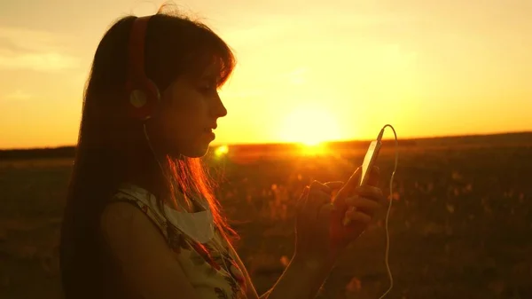 Mädchen, das Musik hört und in den Strahlen eines schönen Sonnenuntergangs tanzt. Ein junges Mädchen mit Kopfhörern und einem Smartphone, das mit dem Finger auf den Sensor des Tablets tippt, wählt online Lieder aus. Zeitlupe. — Stockfoto
