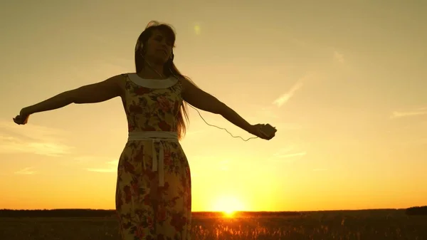 Gelukkig meisje luisteren naar muziek en dansen in stralen van een prachtige zonsondergang tegen de hemel. jong meisje in de hoofdtelefoon en met een smartphone werveling in de vlucht onder de stralen van een warme zonsondergang. Slow Motion. — Stockfoto