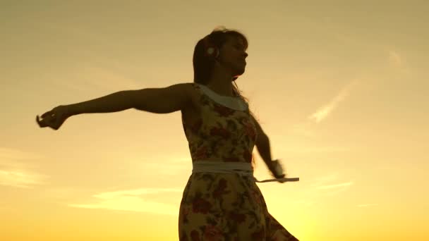 Sexig tjej lyssnar på musik och dans i strålar av en vacker solnedgång mot himlen. ung flicka i hörlurar och med en smartphone virvel i flykt under strålar av en varm solnedgång. Slow motion. — Stockvideo