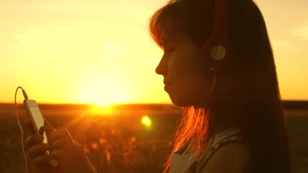 Chica viajero escuchar música en el teléfono inteligente en los rayos de una hermosa puesta de sol de primavera. chica joven con auriculares y con tableta selecciona canciones en línea, en el parque en el verano en los rayos de sol . — Vídeo de stock