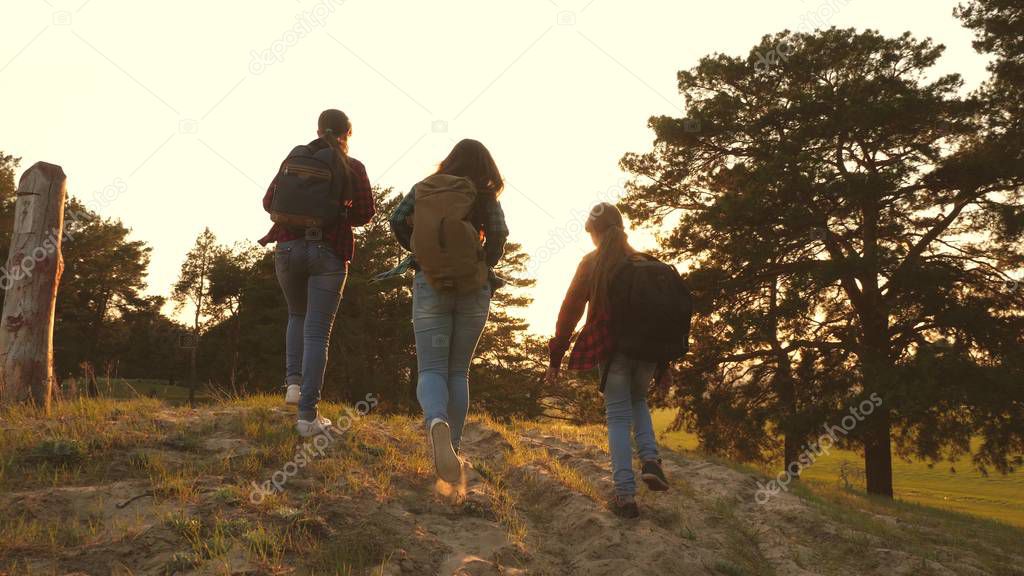 Hiker Girl. three girls travel, walk through woods to climb hill rejoice and raise their hands to the top. girls travel with backpacks on country road. Happy family on vacation travels.