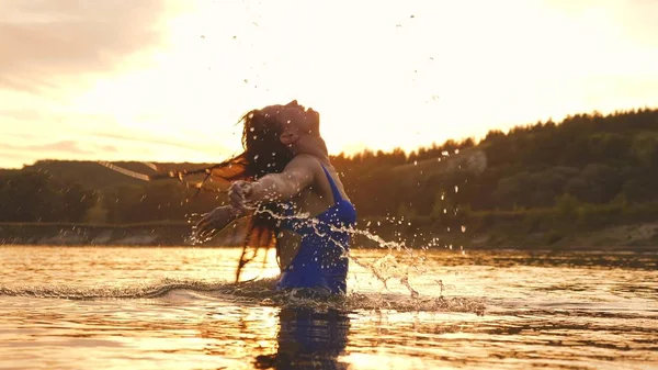 playful girl splashes her long hair in a refreshing summer evening near tropical island with a refreshing river water. girl flips back hair. beautiful spray of water in rays of sunset. splash of water