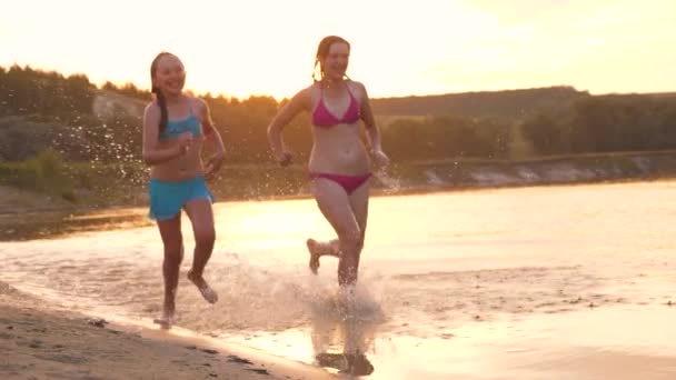 Las niñas corren en la arena a lo largo de la orilla de la playa salpicando gotas de agua y riendo. Los adolescentes felices y libres en las vacaciones veraniegas descansan al atardecer. Trabajo en equipo — Vídeo de stock