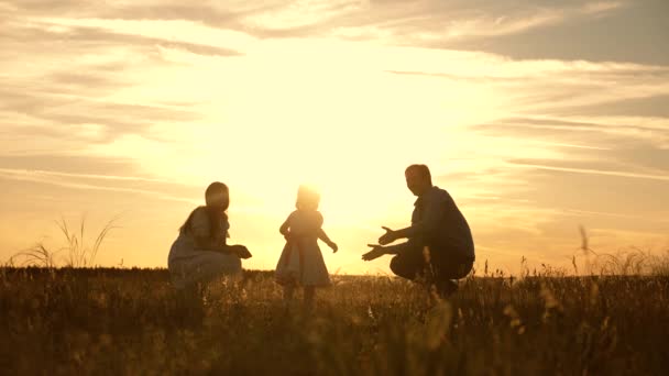 Mamãe e papai brincam com sua filhinha no parque ao pôr do sol, criança dá os primeiros passos. família brincando com sua filha em raios de sol. O bebé vai de pai para mãe e ri. Movimento lento . — Vídeo de Stock