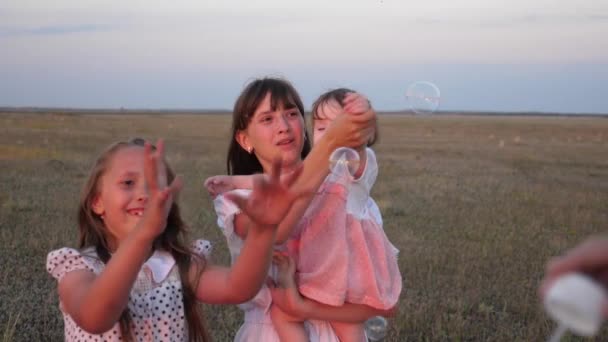 Boldog anya játszik a gyerekekkel fúj szappanbuborékok. gyermek elkapni buborékok és nevetnek a parkban nyáron. Lassított. A koncepció egy boldog család. gyermek, Nővérek és anya együtt játszanak. — Stock videók
