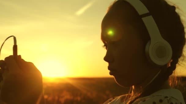 Девочка мечтает под музыку на солнце. Девочка-подросток слушает музыку и смотрит на закат. Счастливая девушка танцует в наушниках в лучах прекрасного восхода солнца в парке . — стоковое видео