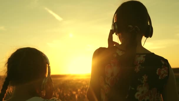 Lyckliga tonårs flickor lyssnar på musik och dansar i strålar av vacker solnedgång i parken. barn i hörlurar under flygning under strålar av en varm solnedgång. systrar lyssnar på musik tillsammans — Stockvideo
