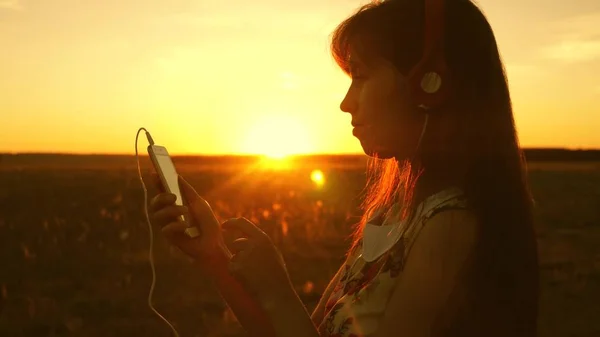 Mädchen, das Musik hört und in den Strahlen eines schönen Sonnenuntergangs tanzt. Ein junges Mädchen mit Kopfhörern und einem Smartphone, das mit dem Finger auf den Sensor des Tablets tippt, wählt online Lieder aus. Zeitlupe. — Stockfoto
