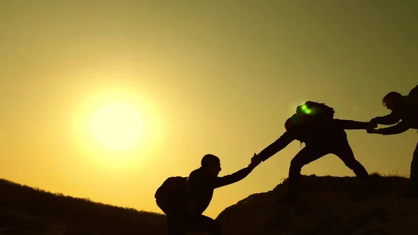 Lavoro di squadra di uomini d'affari. i viaggiatori si arrampicano uno dopo l'altro sulla roccia. affari congiunti. Al rallentatore. Silhouette scalatori allungano le mani l'una all'altra, salendo in cima alla collina . — Foto Stock